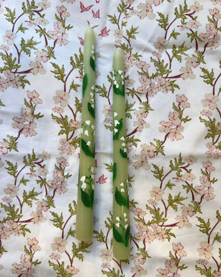 Two light green taper candles decorated with painted green leaves and small white flowers, placed on a white fabric background with a pink and green floral print.