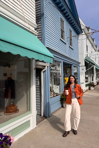 A woman stands on a sidewalk in front of a blue wooden storefront building, wearing an orange jacket, white pants, and dark boots while holding a coffee cup. Storefront awnings in teal and white are visible along the street.