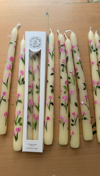 A set of cream-colored taper candles decorated with pink flowers and green leaves, shown both inside and next to their white cardboard packaging box on a wooden tabletop.