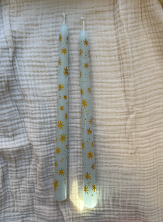 Two white candles with gold star patterns on a textured fabric background